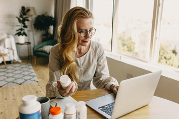 Good-looking freckled ageing woman blogger with long golden hair wearing glasses writing reviews online on new dietary supplements for rejuvenation on her account via social network, using laptop - obrazy, fototapety, plakaty