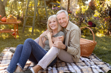 Fototapeta premium Loving elderly couple resting and having picnic in garden, hugging and smiling to camera, sitting together outdoors