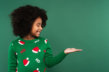 Side view of smiling african american kid in sweater pointing with hand isolated on green
