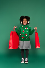 Happy african american child in ear muffs holding shopping bags on green background