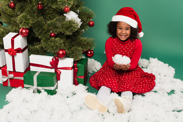 African american kid in santa hat holding decorative snow near presents and christmas tree on green background