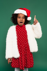 Excited african american girl in fluffy jacket and santa hat having idea isolated on green