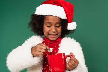 Happy african american child in santa hat and fluffy jacket holding marshmallow near cup isolated on green