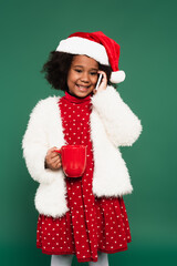 African american girl in santa hat and fluffy jacket talking on smartphone and holding cup isolated on green