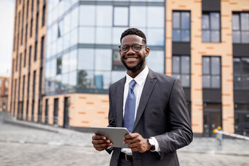 Business news. Happy black businessman using digital tablet walking outdoors near modern office building, copy space