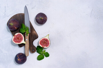 Round ripe figs cut into halves lies on a dark wooden plank. Green mint leaves and a knife in the background