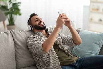 Smiling young Arab guy in headphones listening to music or audio book on couch at home. Weekend pastimes concept