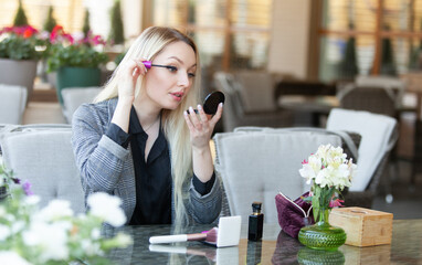 Young attractive beauty woman blonde applying makeup (paint her eyelashes with mascara brush) on the summer terrace of cafe