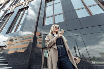 Blonde woman in trench coat and sunglasses uses smartphone in urban location