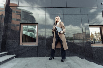 Stylish blonde business woman with laptop in trench coat in urban location