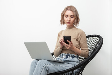 Business serious woman millennial uses a smartphone and sits on an armchair with a laptop against the background of a white wall. Remote work