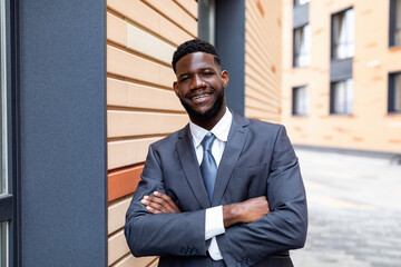 Portrait of confident black businessman in suit posing with crossed arms by office center and smiling at camera
