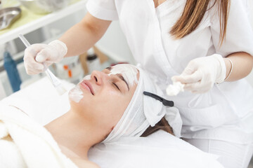 Cosmetological procedures. Beauty salon, doctor in gloves applies mask for beautiful woman with closed eyes with brush.
