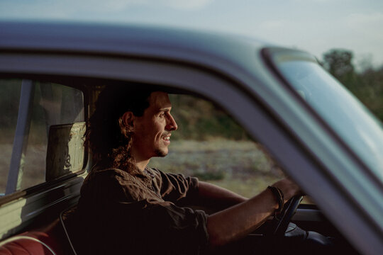 Happy smile confident young handsome man driver with a beard smiling with glad positive expression in the pickup car truck. Attractive male driving vehicle hand on steering wheel. Sunset evening day