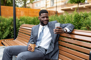 Fototapeta premium Smiling african american manager holding takeaway coffee and using smartphone while sitting on bench outdoors
