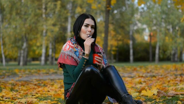 Beautiful stylish young girl portrait brunette drinks hot coffee and smokes an electronic cigarette in the autumn city park