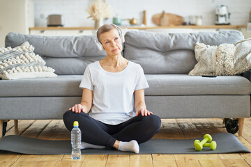 Senior woman sit cross-legged in headphones listen music in living room. Relax after exercising...