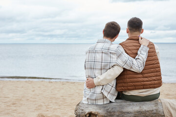 Fototapeta premium Back view portrait of young gay couple embracing on beach while enjoying sea view, copy space