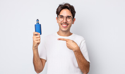 young hispanic man smiling cheerfully, feeling happy and pointing to the side. smoke vaporizer...