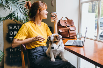 Young girl student sititng at table with computer and dog at work place. Pretty amazing female...