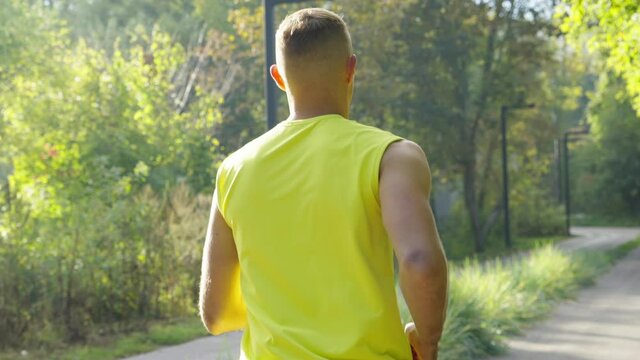 Back View Of Athletic Man With Short Haircut Wearing Yellow Top Running In Park In Slow Motion, Breathing Out Steam. Following Shot Of Runner Training On Chilly Morning. Concept Of Sport