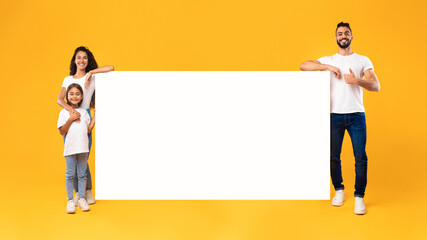 Middle-Eastern Family Standing Near Big Empty Poster On Yellow Background
