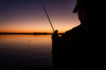 Silhouette of a man fishing on the shore of the river at sunset.