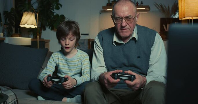 Cute teen boy grandchild teaching happy senior 60s grandfather to play console games with joysticks at home on sofa.