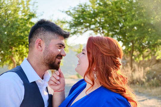 Gorgeous happy caucasian plus size red headed woman touches her husband smiling. Mixed race couple engagement. Middle eastern man and caucasian woman hugging.