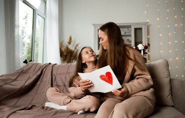 mom and daughter are sitting on the couch and looking at a postcard with a heart