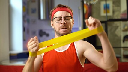 Portrait of funny man in red retro sportswear doing exercises with an elastic resistance band at home.