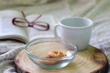 Bowl of cookies, cup, book and reading glasses on a bed. Selective focus.