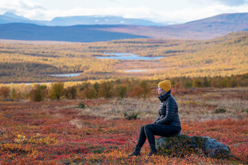 Caucasian outdoor active young adult woman sitting on Rock With outdoor hiking cloths jacket and cap, looking away from camera with Autumn colored floorbed and mountains in pieljekaise national park.