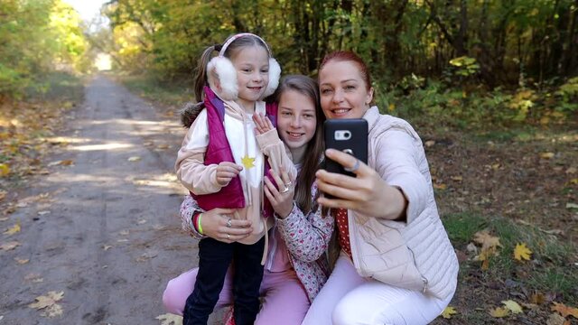 a cheerful woman takes selfies with her daughters in an autumn park, family photos of happy people