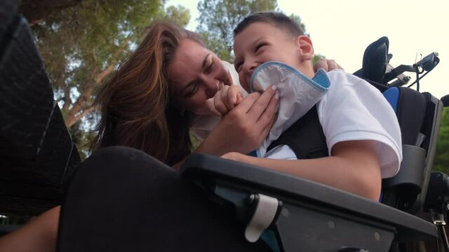 Loving mother feeling bond and love playing kissing on cheek her little 9s son with cerebral palsy sit in wheelchair outdoor