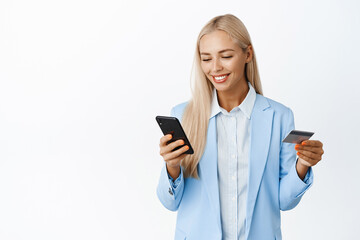 Smiling blond corporate woman enter info on mobile phone, holding credit card, wearing suit,...