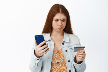 Confused young redhead girl holding mobile phone, looking troubled at credit card, having problem with online order, unable to make purchase, white background