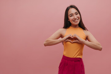Beautiful asian young woman smiling teeth making heart with hands while standing on pink background. Image of happy teenager with black long hair wearing yellow top and raspberry pants. 