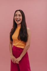 Laughing young 20s asian girl wearing orange top and crimson pants on pink background. Side view image of long and dark-haired woman holding hands together. Summer playful mood concept