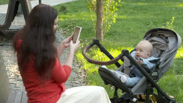 Mother photographing baby in stroller with smartphone outdoors, cute caucasian six month old child. High quality 4k footage