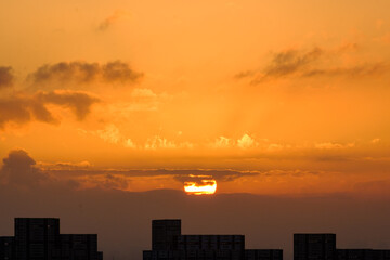  都市の夜明け、早朝ビルの隙間から太陽が昇り辺りはオレンジ色に染まる。ビルはシルエットに浮かぶ