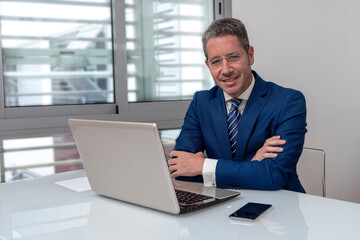 CAUCASIAN BUSINESSMAN WITH FOLDED ARMS AT MODERN OFFICE WITH LAPTOP SMILING AND LOOKING AT CAMERA. CONCEPT OF EXECUTIVE, MANAGER, BOSS, DIRECTOR OR ENTREPENEUR.