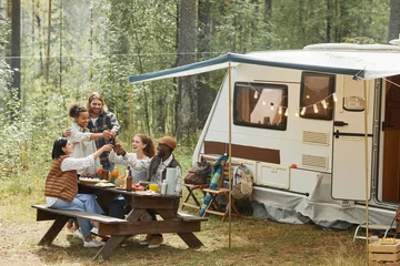 Poster Brede kijkhoek op diverse groepen jongeren die bierflesjes rammelen terwijl ze genieten van een picknick buiten op de camping met een aanhangwagen © Seventyfour