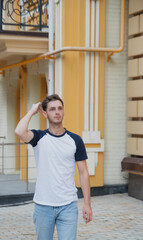 A boy with brown hair in white t-shirt is walking on the street with beautiful architecture on the back. White t-shirt with copy space for text or picture. Stylish man on the street photoshoot.