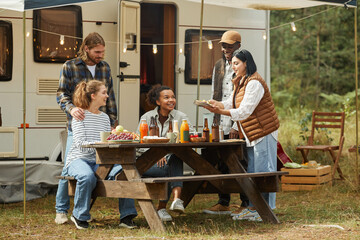Full length view at diverse group of young people enjoying picnic outdoors while camping with trailer van - obrazy, fototapety, plakaty