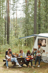 Vertical wide angle view at diverse group of young people enjoying picnic outdoors while camping with trailer van, copy space
