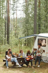 Fotobehang Verticale brede kijkhoek bij diverse groepen jongeren die buiten genieten van een picknick terwijl ze kamperen met een aanhangwagen, kopieer ruimte © Seventyfour