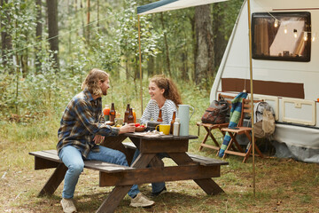 Full length shot of young couple enjoying picnic outdoors while camping with trailer van, copy space