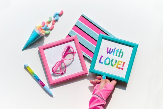 Text With love in picture frame in hand. Sweets and vibrant party decor objects. Candy, snacks, funny heart shaped gasses in picture frames. Festive objects on white background. Sunlight, shadows.
