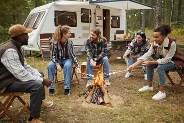 Diverse group of young people roasting marshmallows while enjoying camping with friends in forest,...